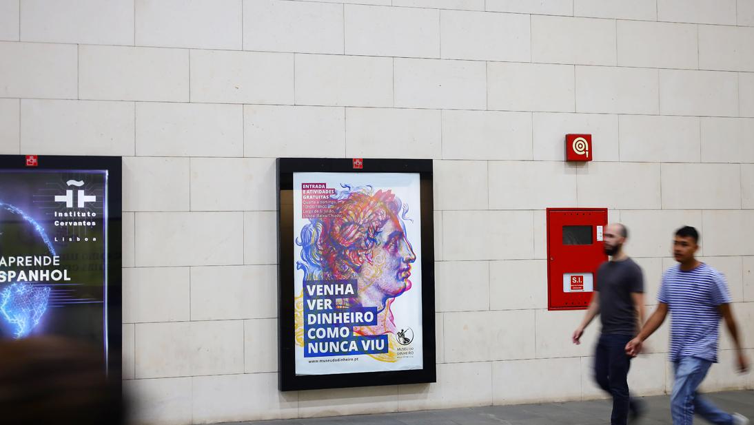 Pessoas caminham no metro de Lisboa e passam perto de um mupi com um cartaz do Museu do Dinheiro